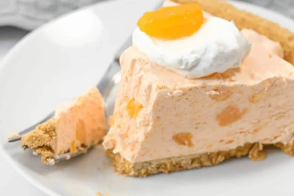 A slice of creamy orange dessert pie with a graham cracker crust, topped with whipped cream and an orange segment, on a white plate with a fork—perfect for anyone who loves easy pie recipes.