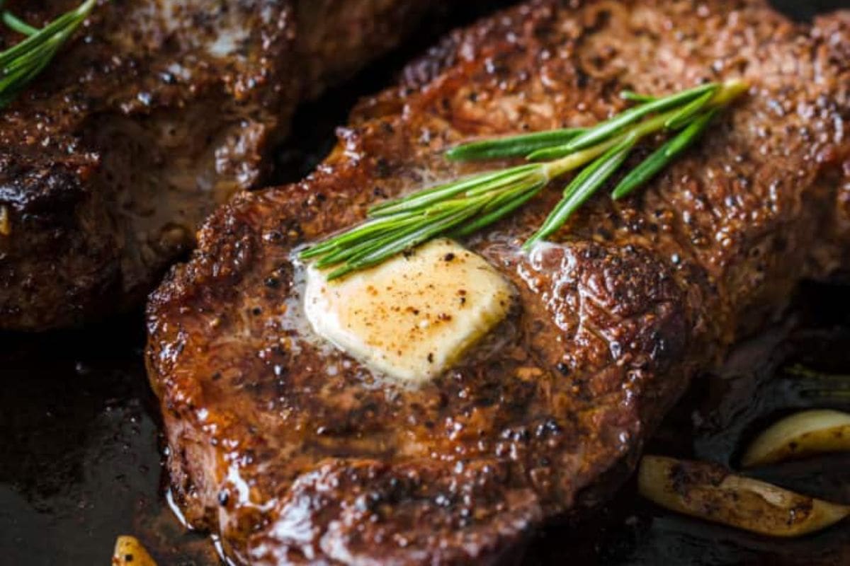 A juicy steak topped with a melted pat of butter and a sprig of rosemary, with visible seasoning and seared edges—an irresistible treat for fans of classic steakhouse flavors and steak recipes.