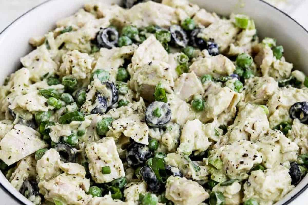 A bowl of creamy chicken salad mixed with green peas, black olives, chopped green onions, and diced chicken, seasoned with black pepper and a touch of bold pesto for extra flavor. Perfect for fans of flavorful pesto recipes.