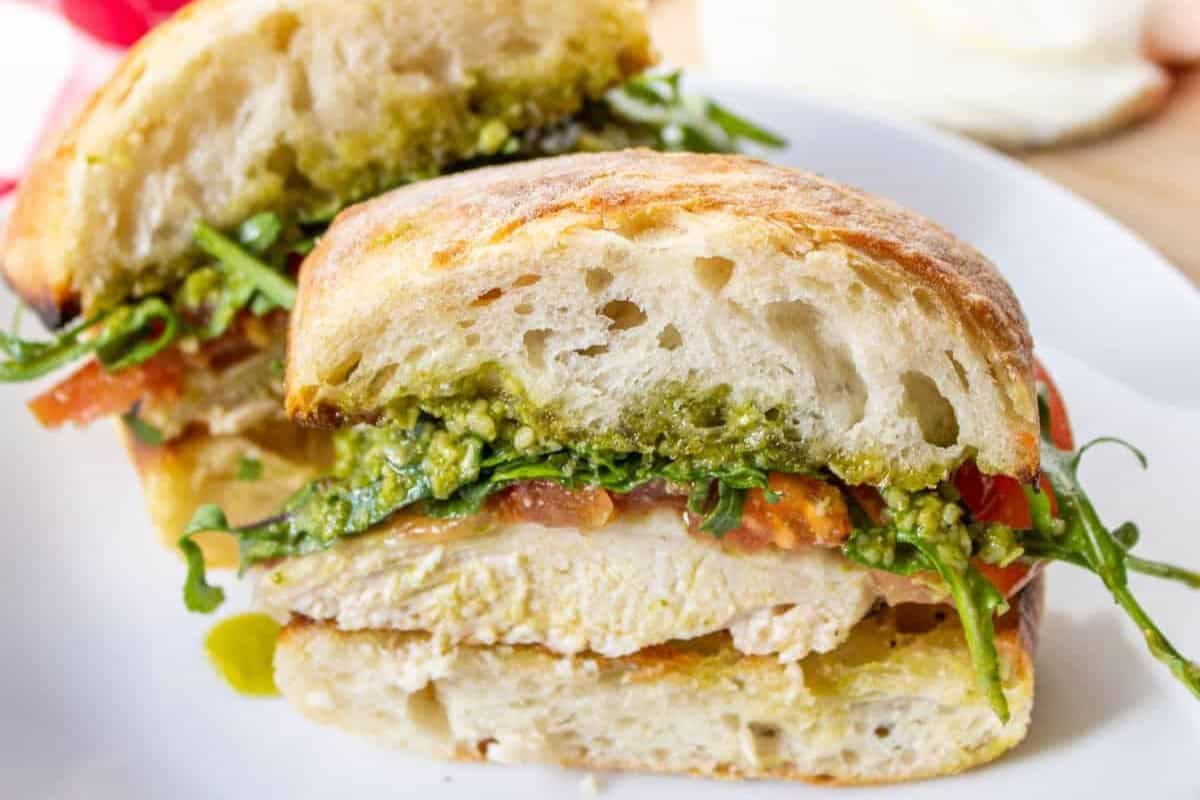 A sliced chicken sandwich on ciabatta bread with bold pesto, arugula, tomato, and cheese on a white plate.