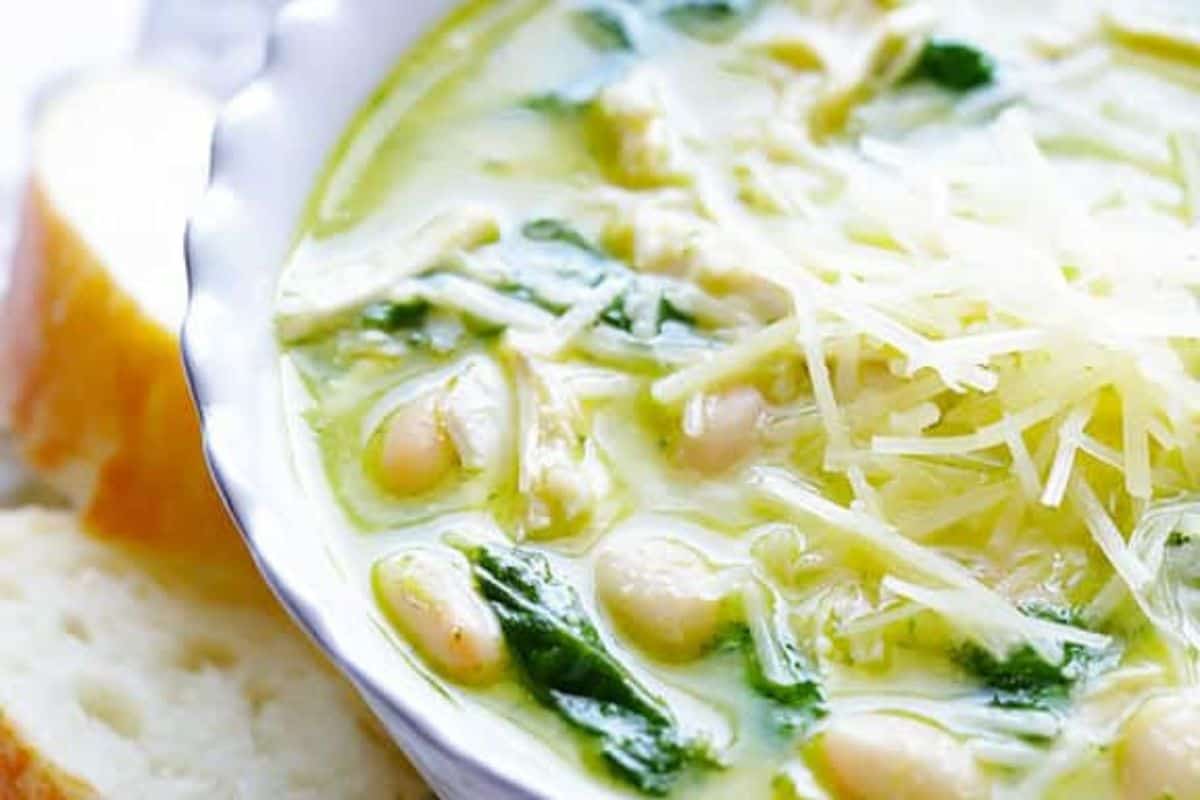 A bowl of creamy chicken and pesto soup topped with shredded cheese and a swirl of bold pesto, served with slices of bread on the side.