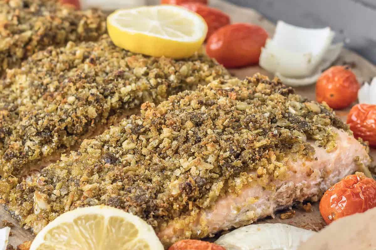 Close-up of baked salmon fillets with a herb and nut crust, topped with bold pesto and garnished with lemon slices, cherry tomatoes, and onion pieces.