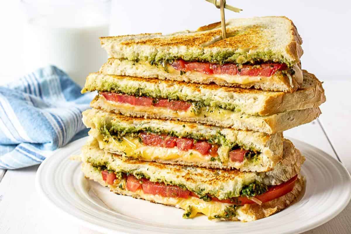 Three grilled cheese and tomato sandwiches with bold pesto are stacked on a white plate, held together with a toothpick. A glass of milk and a cloth napkin are in the background—perfect for fans of flavorful pesto recipes.