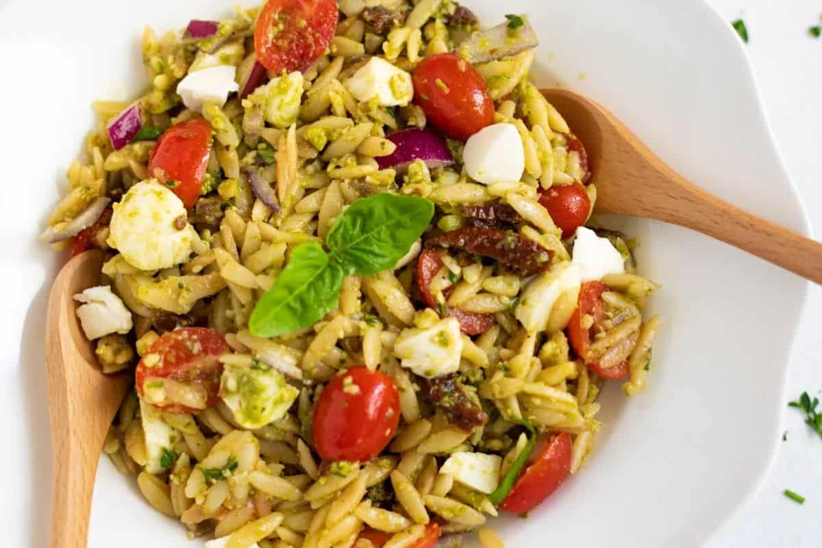 A bowl of orzo pasta salad with cherry tomatoes, mozzarella, red onions, sun-dried tomatoes, basil, and a wooden spoon—perfect for pairing with bold pesto or exploring new pesto recipes.