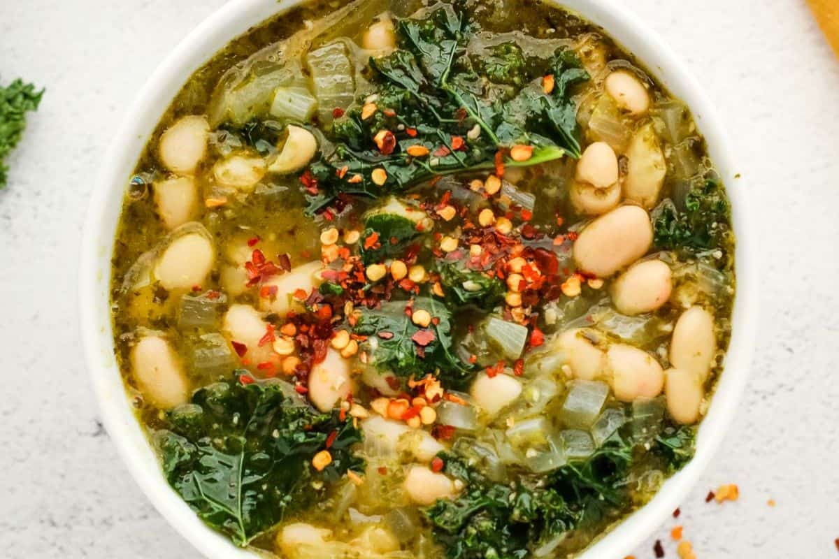 A bowl of soup with white beans, chopped kale, onions, and red pepper flakes on top, finished with a swirl of bold pesto for extra flavor, set on a light surface.