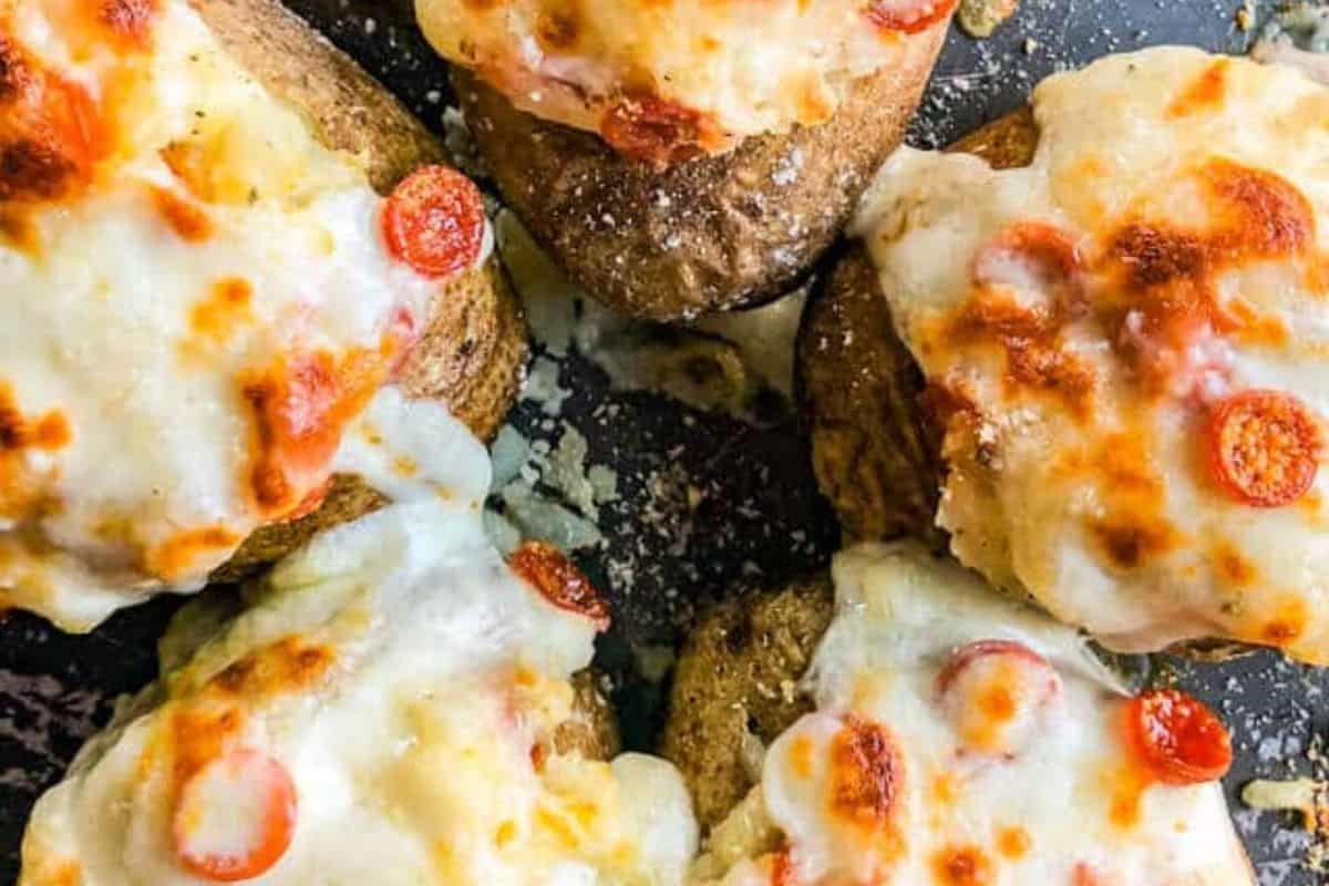 Five baked potatoes topped with melted cheese and small pepperoni slices are arranged closely together on a dark surface, making this a tasty addition to your list of creative baked potato ideas.