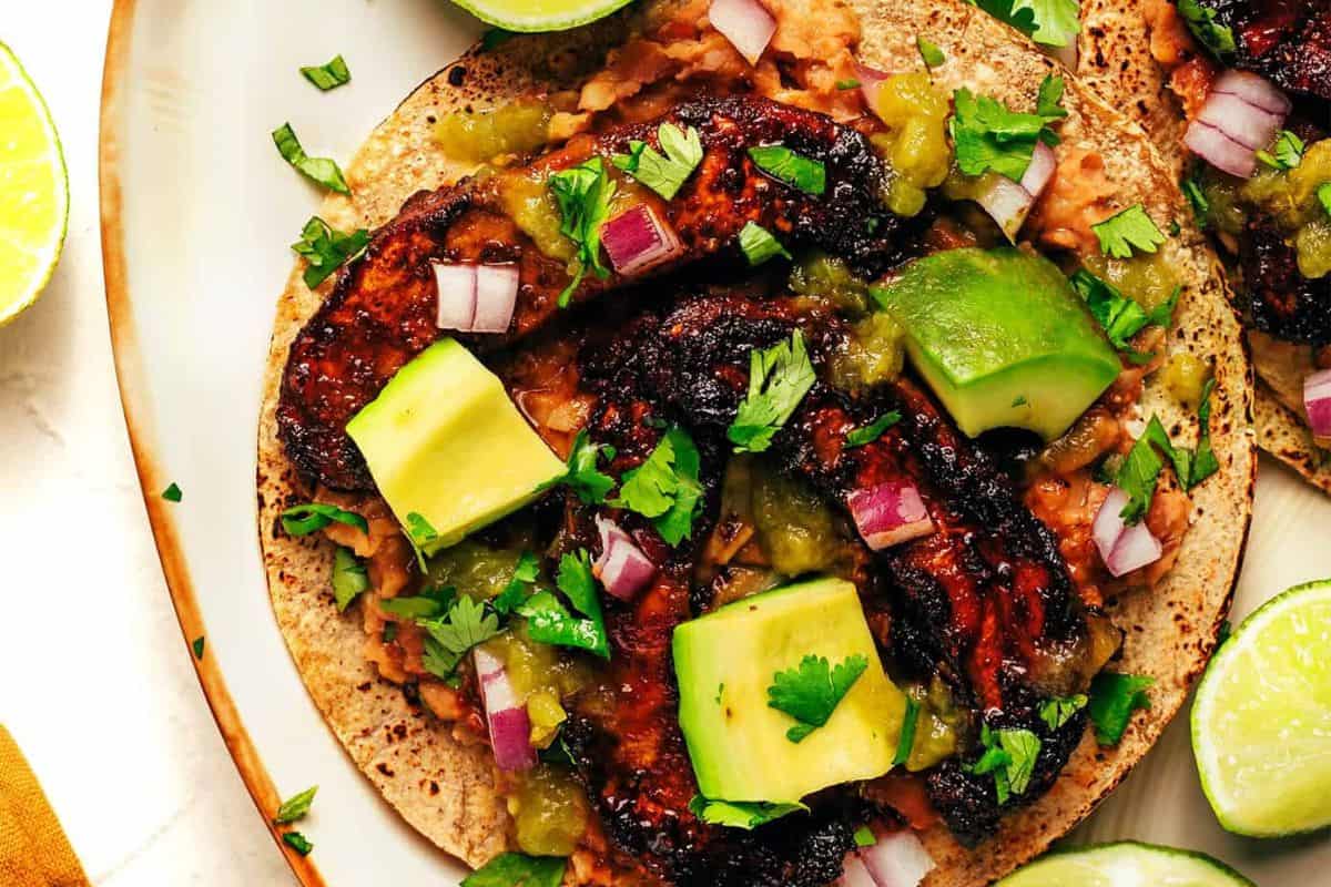 A must-try close-up of a taco with mushrooms, avocado cubes, chopped red onion, cilantro, green salsa, and refried beans on a corn tortilla, garnished with lime wedges—taking taco night beyond the basics.