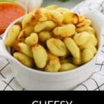 A white bowl filled with Cheesy Party Potatoes sits on a cloth, surrounded by parsley, garlic, and a cup of red sauce. Text reads: "Cheesy Party Potatoes.