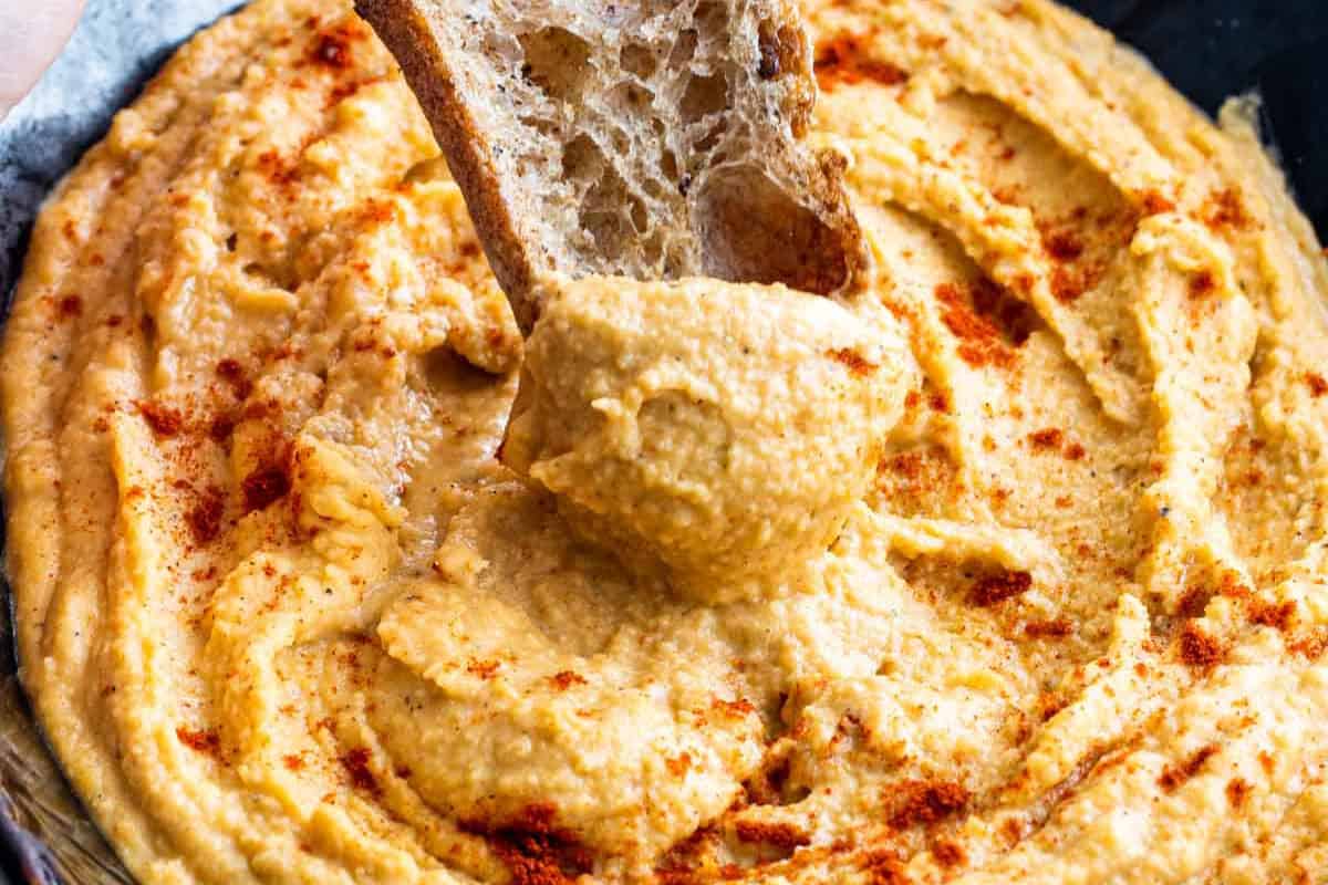 A slice of bread is dipped into a bowl of delicious hummus topped with a sprinkle of paprika—a perfect example of how simple hummus recipes can elevate any snack.
