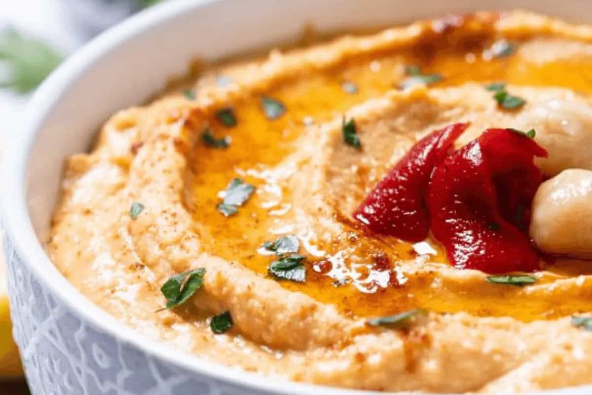A bowl of classic hummus garnished with olive oil, parsley, whole chickpeas, and roasted red pepper—one of the delicious twists on traditional hummus recipes.