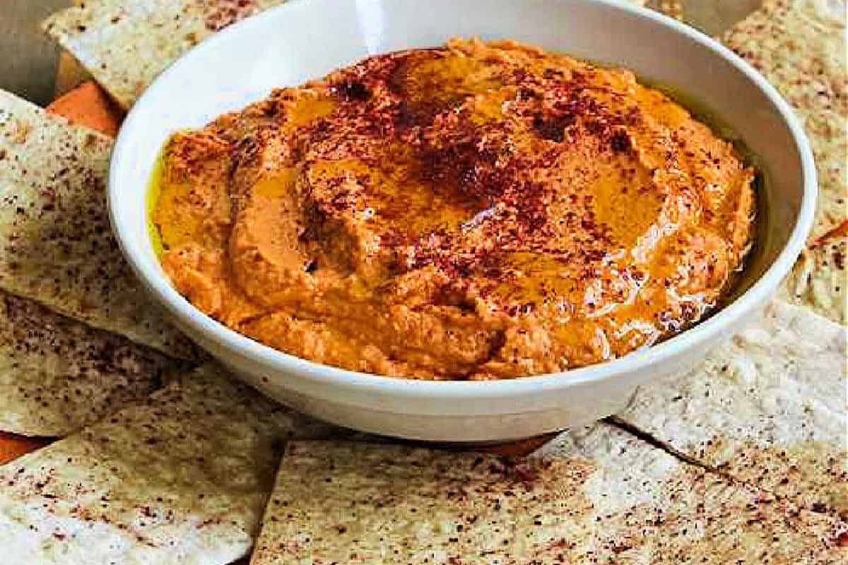 A white bowl filled with delicious hummus made from red pepper, drizzled with olive oil and sprinkled with paprika, surrounded by pieces of flatbread.
