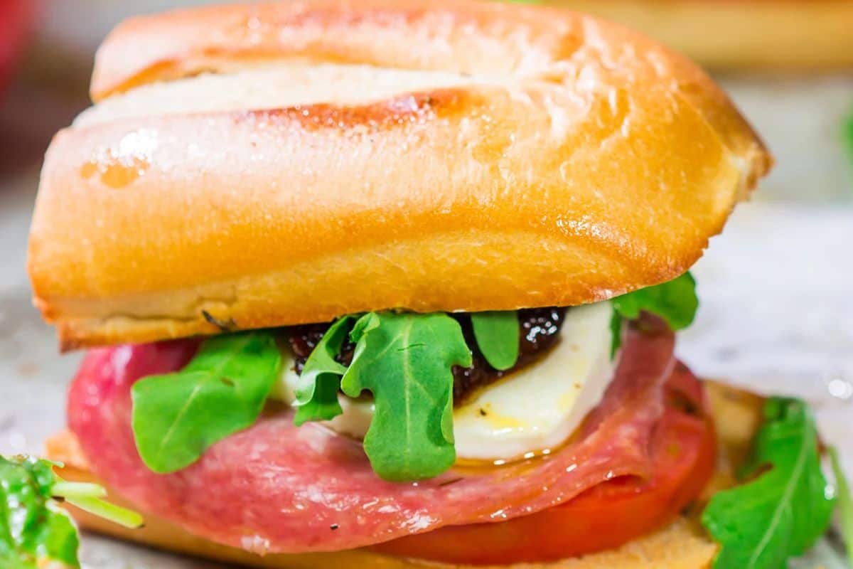 A close-up of a sandwich with salami, tomato, arugula, cheese, and a bold pesto spread on a crusty baguette.