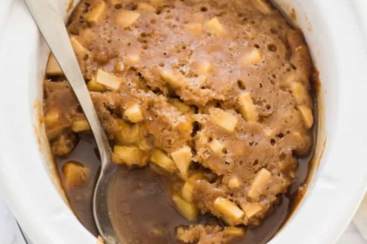 A baked apple pudding dessert with chunks of apple and a caramel-like sauce, served in a white baking dish—an effortless indulgence perfect for fans of slow cooker desserts. A serving spoon rests inside, ready to enjoy.