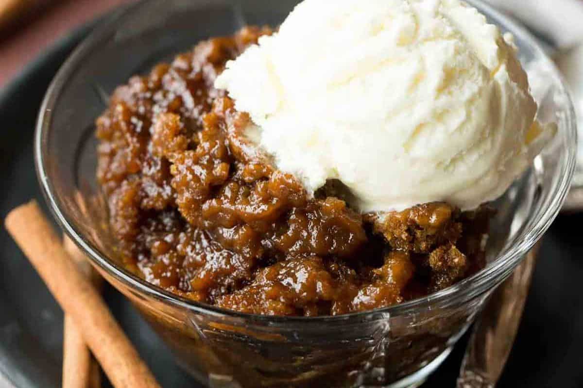 A glass bowl with sticky toffee pudding topped with a scoop of vanilla ice cream, next to two cinnamon sticks—an inviting treat that showcases the magic of Slow Cooker Desserts.