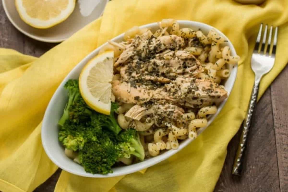 Oval white bowl with pasta, shredded chicken, herbs, broccoli, and a lemon wedge on a yellow cloth—this dish is finished with bold pesto for extra zest, with a fork and plate in the background.