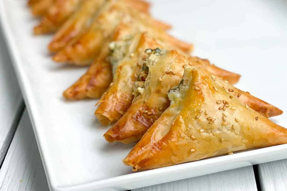 A row of golden, triangular pastries topped with sesame seeds is arranged neatly on a white rectangular plate—perfect for those seeking elegant cold appetizer ideas.