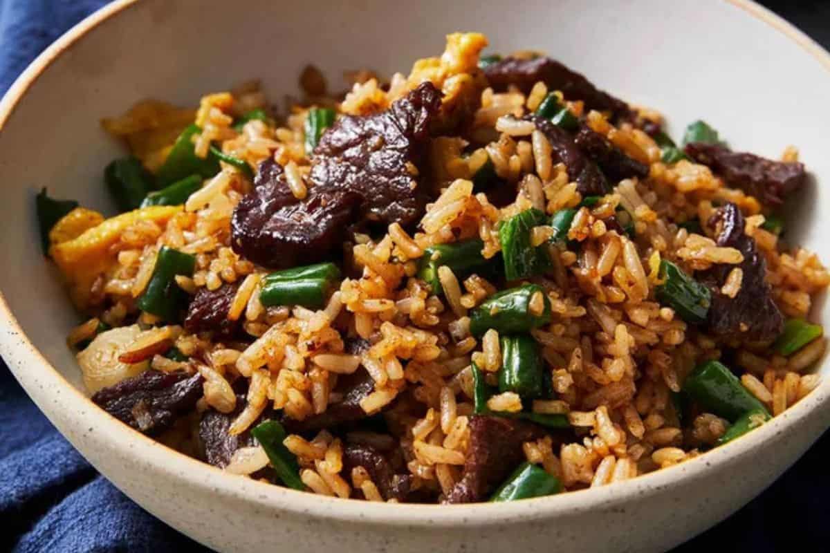 A bowl of beef fried rice with green beans, egg, and scallions sits on a dark blue cloth background—perfect for fans of juicy steak and classic steakhouse flavors.