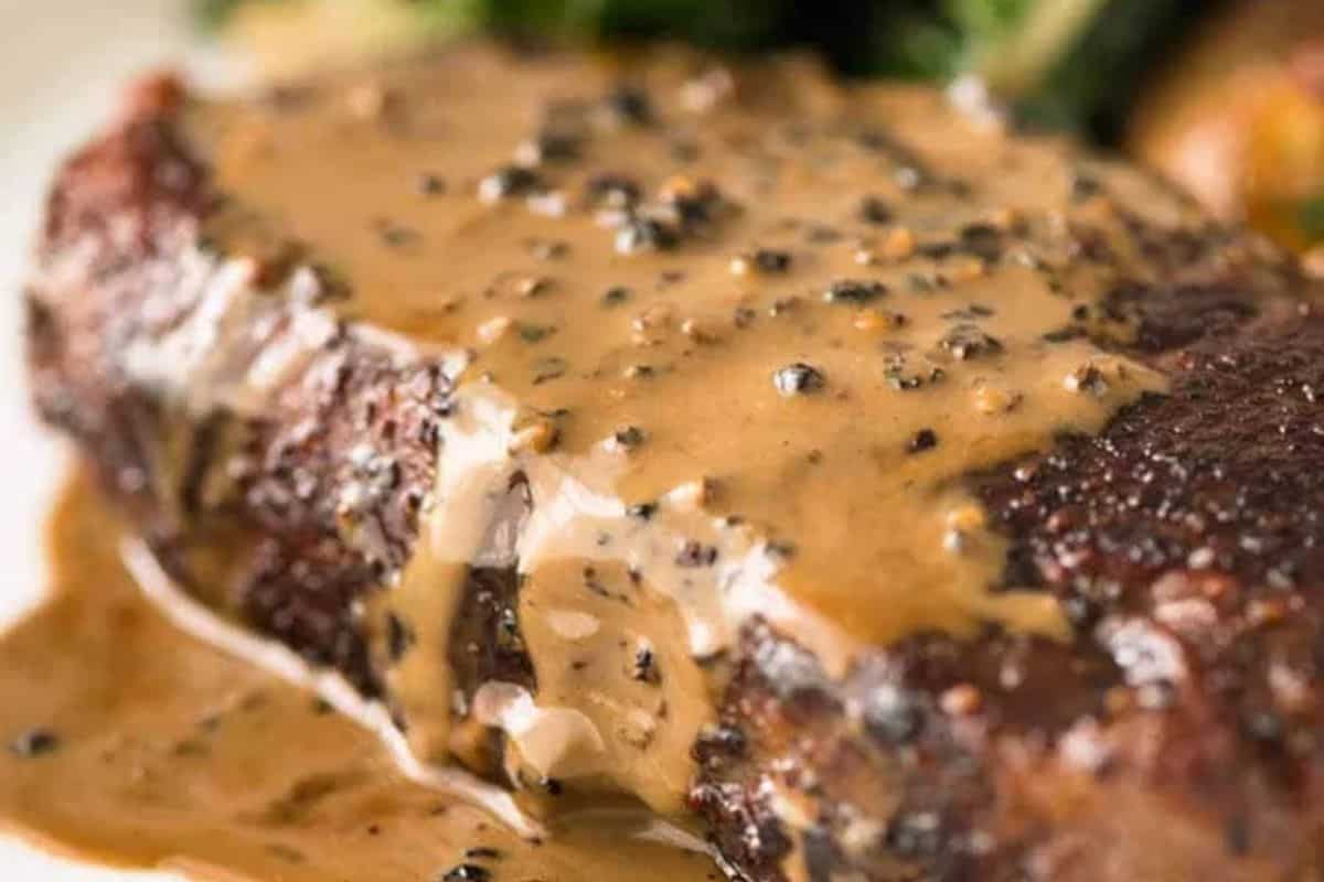 A close-up of a juicy steak topped with creamy peppercorn sauce, with some vegetables visible in the blurred background—perfect inspiration for your next steakhouse-worthy steak recipes.