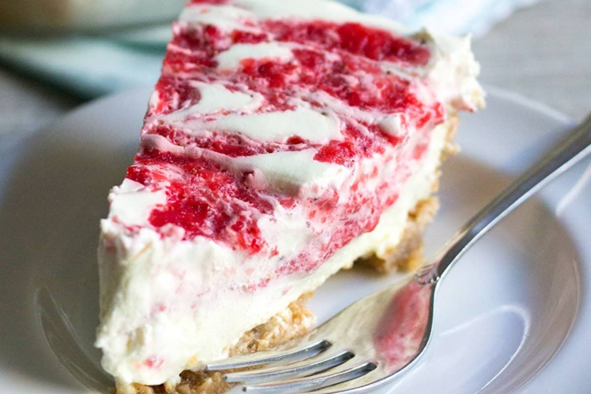 A slice of strawberry swirl cheesecake with a crumbly crust sits on a white plate with a fork—perfect inspiration for those seeking easy pie recipes.