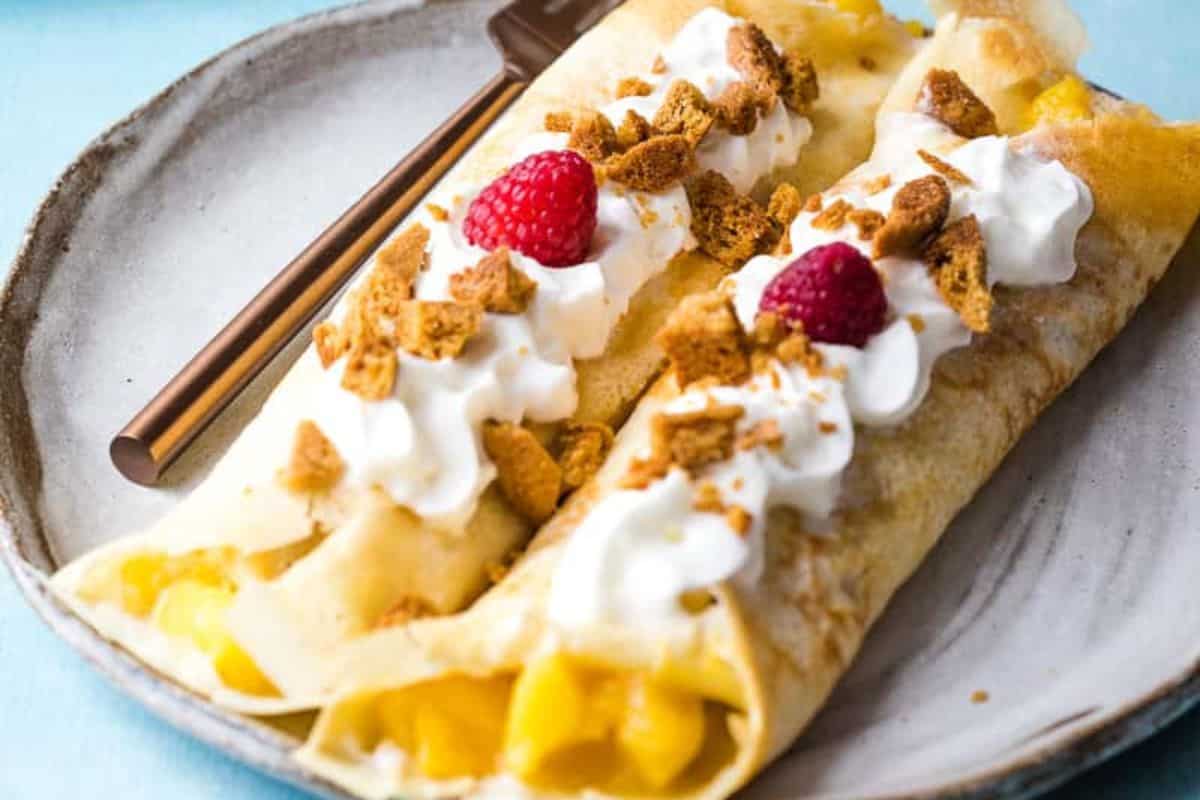 A close-up of two sweet mango crepes, topped with wipped cream, graham crackers, and raspberries.
