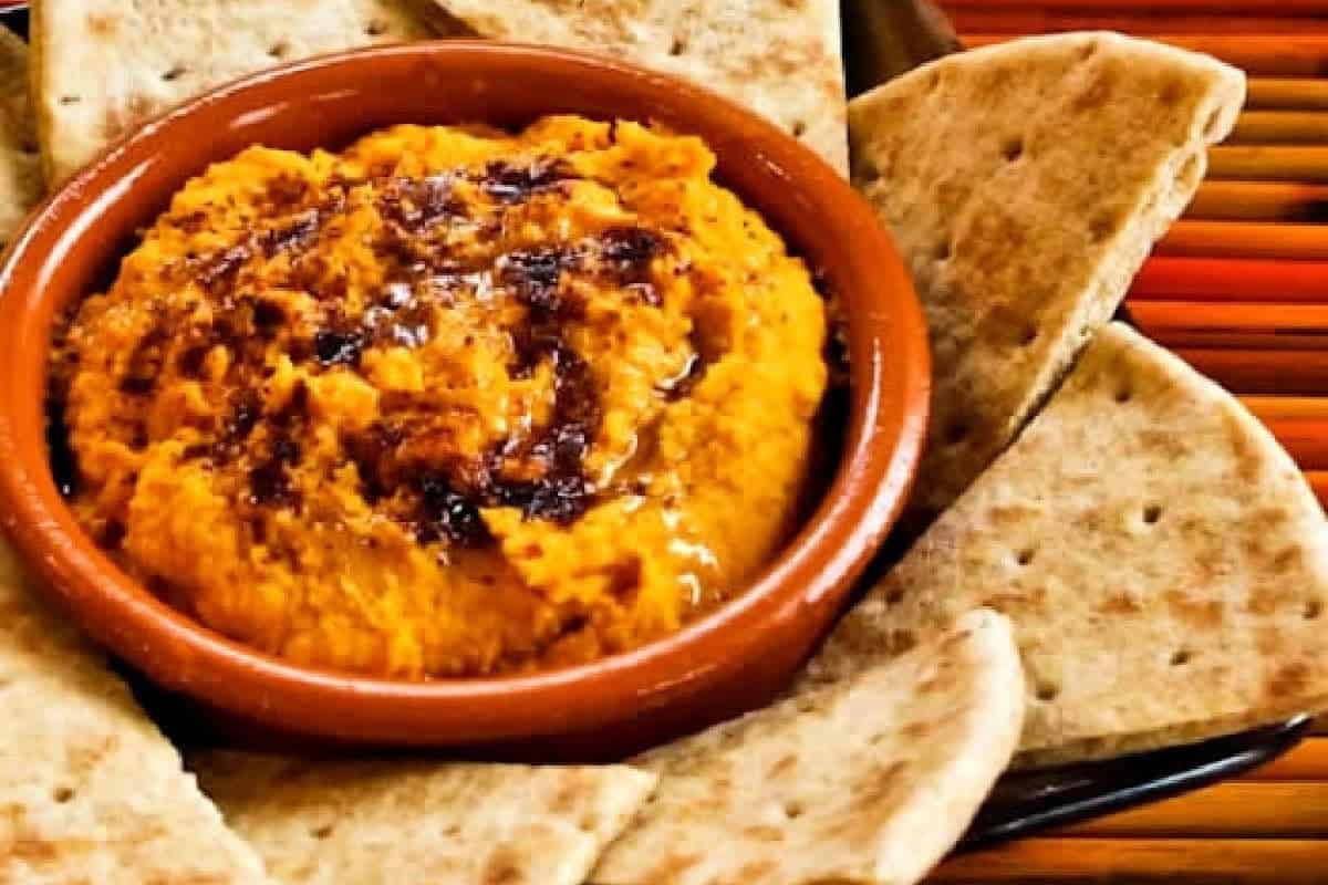 A bowl of orange-colored dip, possibly Classic Hummus with Delicious Twists, garnished with spices and surrounded by pieces of pita bread.