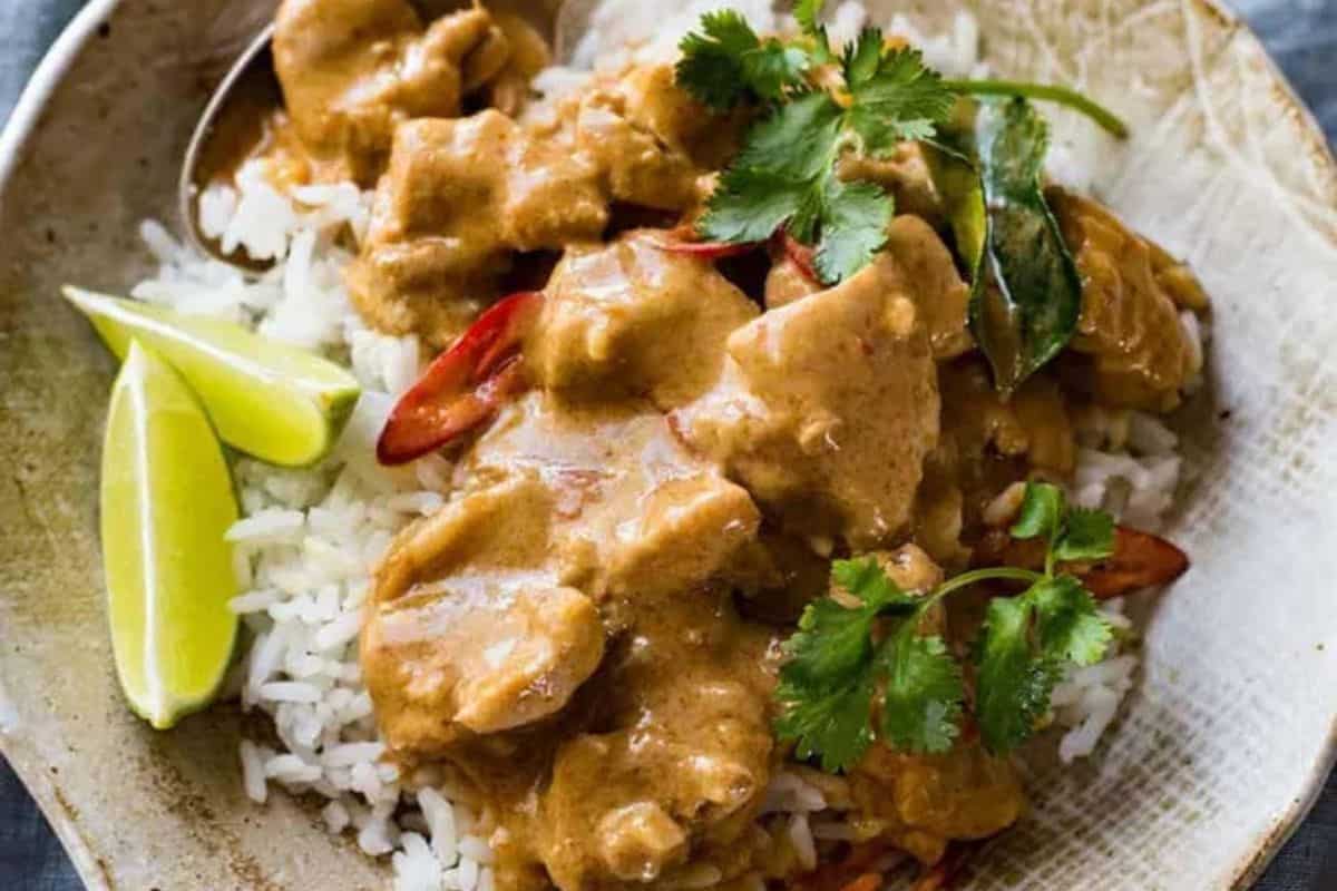 A close-up of a bowl of thai mango chicken curry, served over rice with lime wedges on the side.