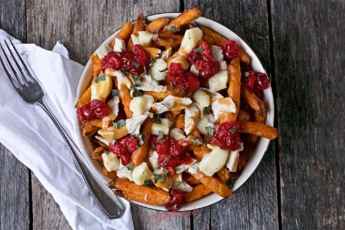 A plate of sweet potato fries bursting with flavor, topped with melted cheese, shredded turkey, and cranberry sauce, placed on a wooden surface with a fork and napkin beside it.