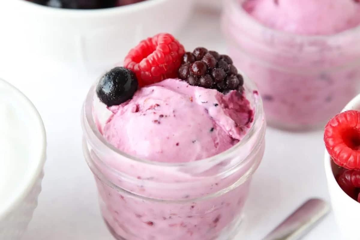 A small glass jar filled with purple berry ice cream, one of summer’s irresistible desserts, topped with a raspberry, blackberry, and blueberry, with more jars and bowls of frozen desserts in the background.