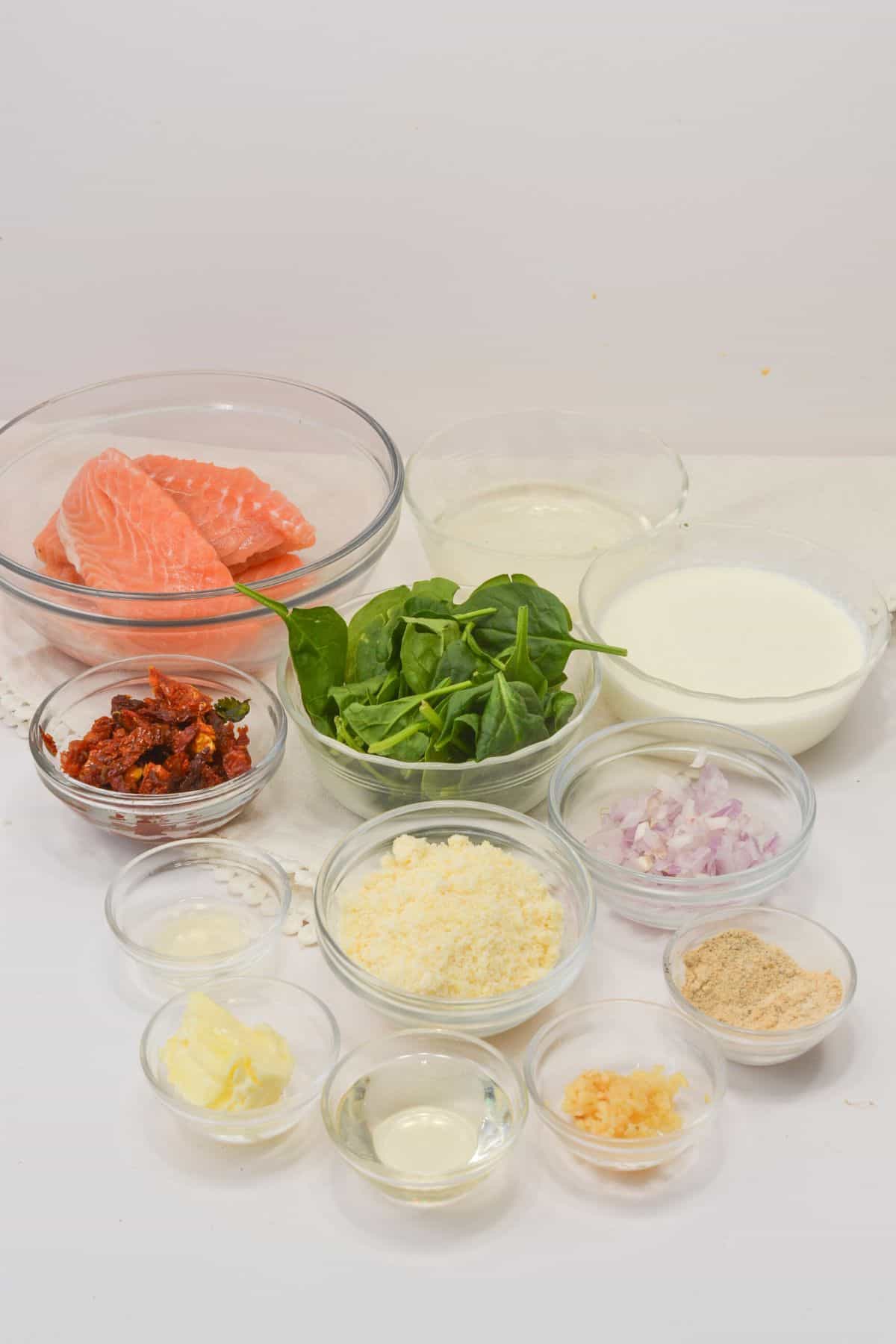 A variety of ingredients in glass bowls for including raw fish, spinach, sun-dried tomatoes, cream, Parmesan cheese, chopped shallots, garlic, butter, breadcrumbs, and oil.