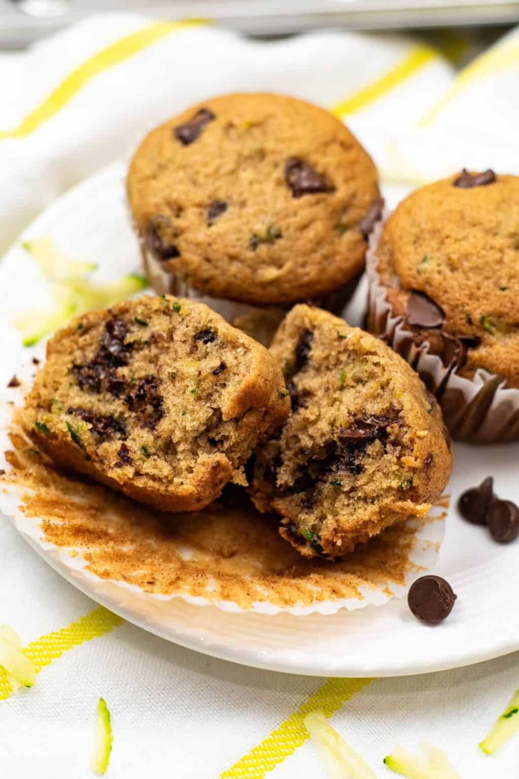 Three Zucchini Chocolate Chip Muffins sit on a white plate, with one split open to reveal its moist interior filled with chocolate chips and green zucchini pieces.
