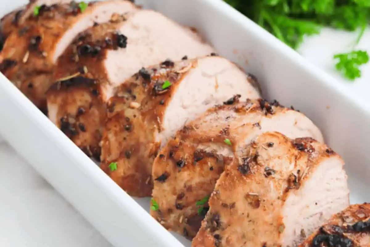 Sliced, seasoned pork tenderloin arranged in a white serving dish with fresh herbs in the background—a perfect addition to your collection of delicious pork recipes.