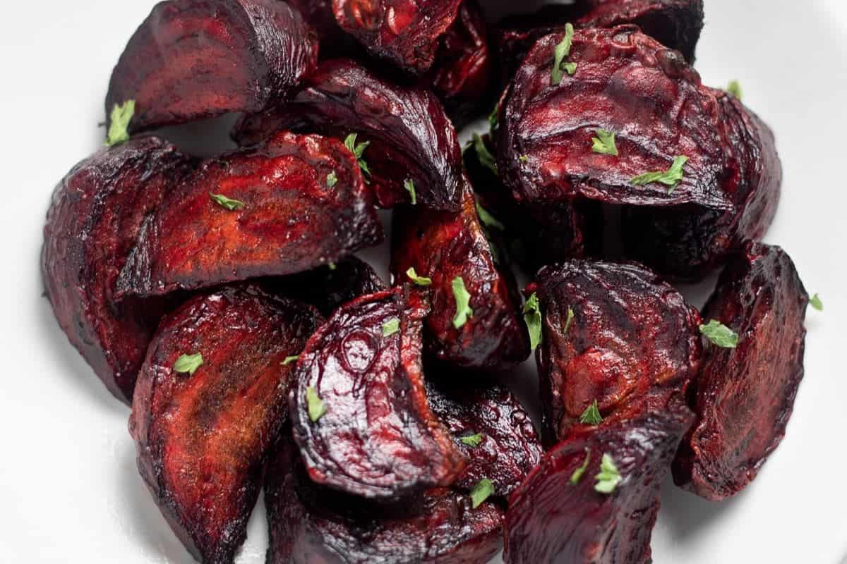 A close-up of roasted beets, cut into rustic chunks, with slightly browned edges and sprinkled with fresh green herbs, presented on a white background.