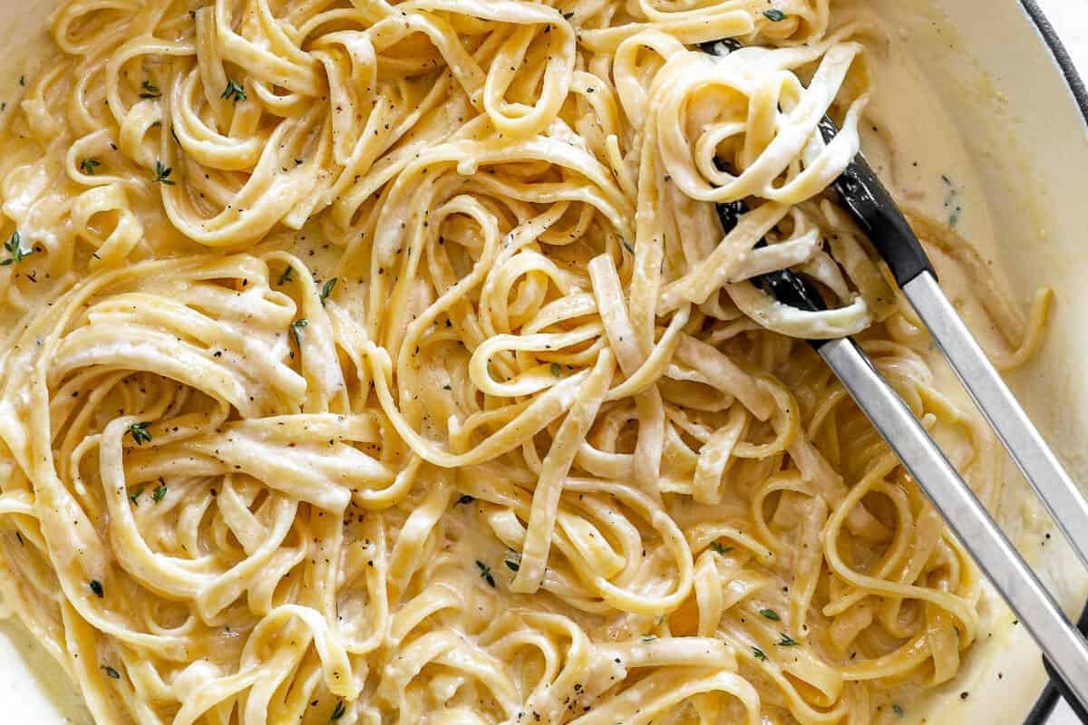 Tongs serving creamy fettuccine Alfredo pasta with a light sprinkling of black pepper and herbs—a comfort dish inspired by hearty recipes perfect for any Dutch oven enthusiast.