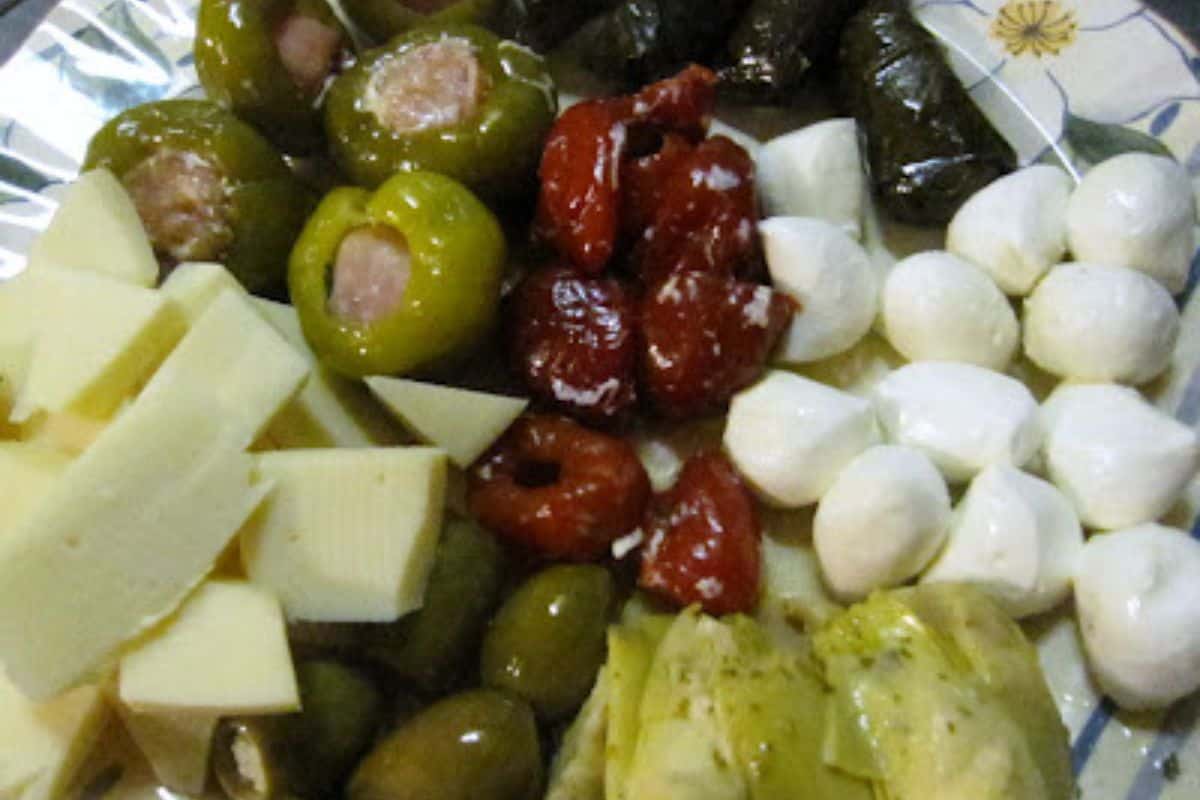  A close-up of an antipasto platter featuring stuffed green peppers, red sun-dried tomatoes, small white mozzarella balls, cut white cheese, green olives, and artichoke hearts.