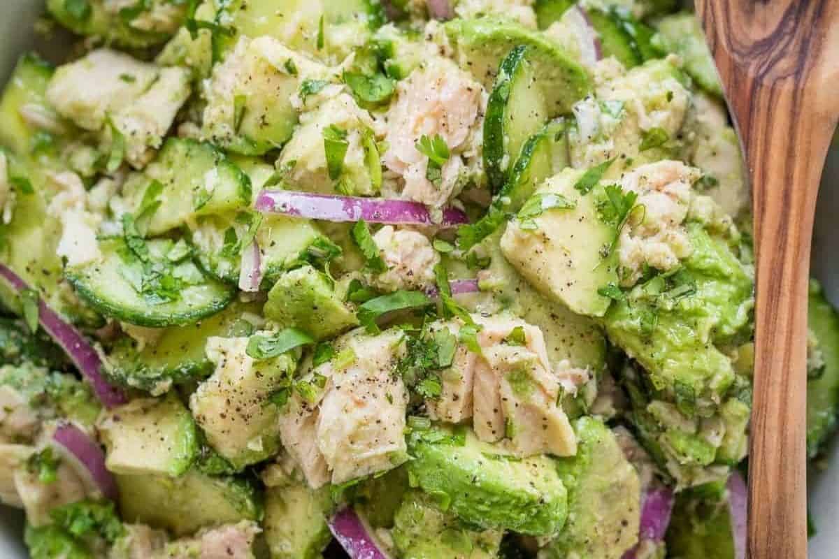 A close-up of a salad with chunks of canned tuna, avocado, sliced cucumber, red onion, and fresh herbs, seasoned with black pepper. A wooden spoon is partially visible on the side—perfect for those seeking easy dinner ideas.