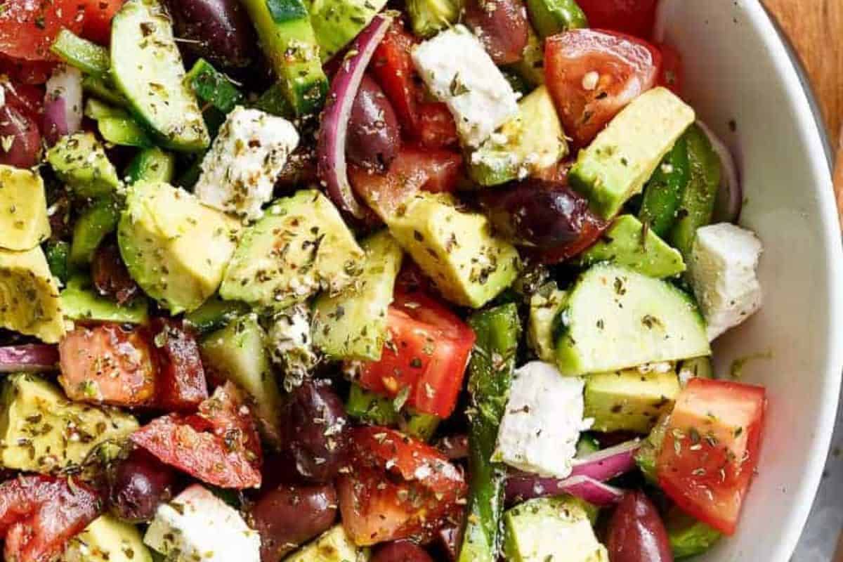 A vibrant Greek salad with large chunks of avocado, cucumber, tomatoes, red onion, black olives, and feta cheese, all sprinkled with herbs.