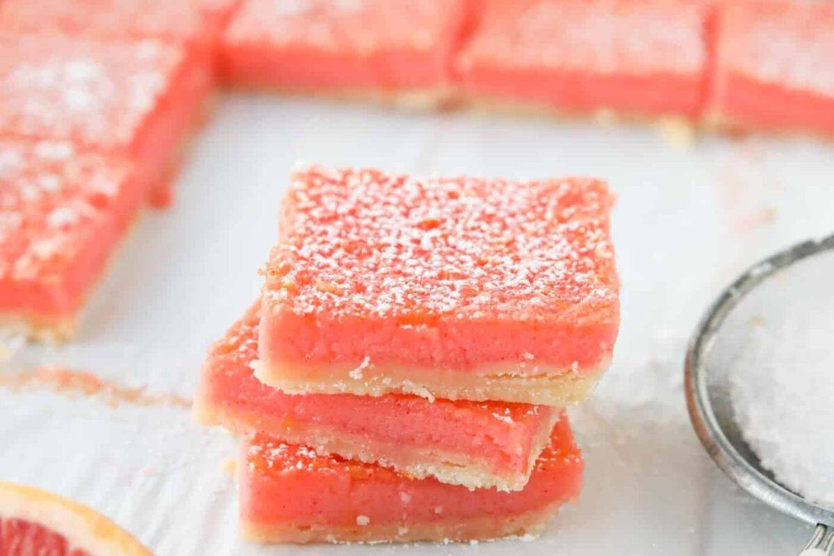 Three pink dessert bars dusted with powdered sugar are stacked on a white surface, with more bars and a sifter visible in the background.