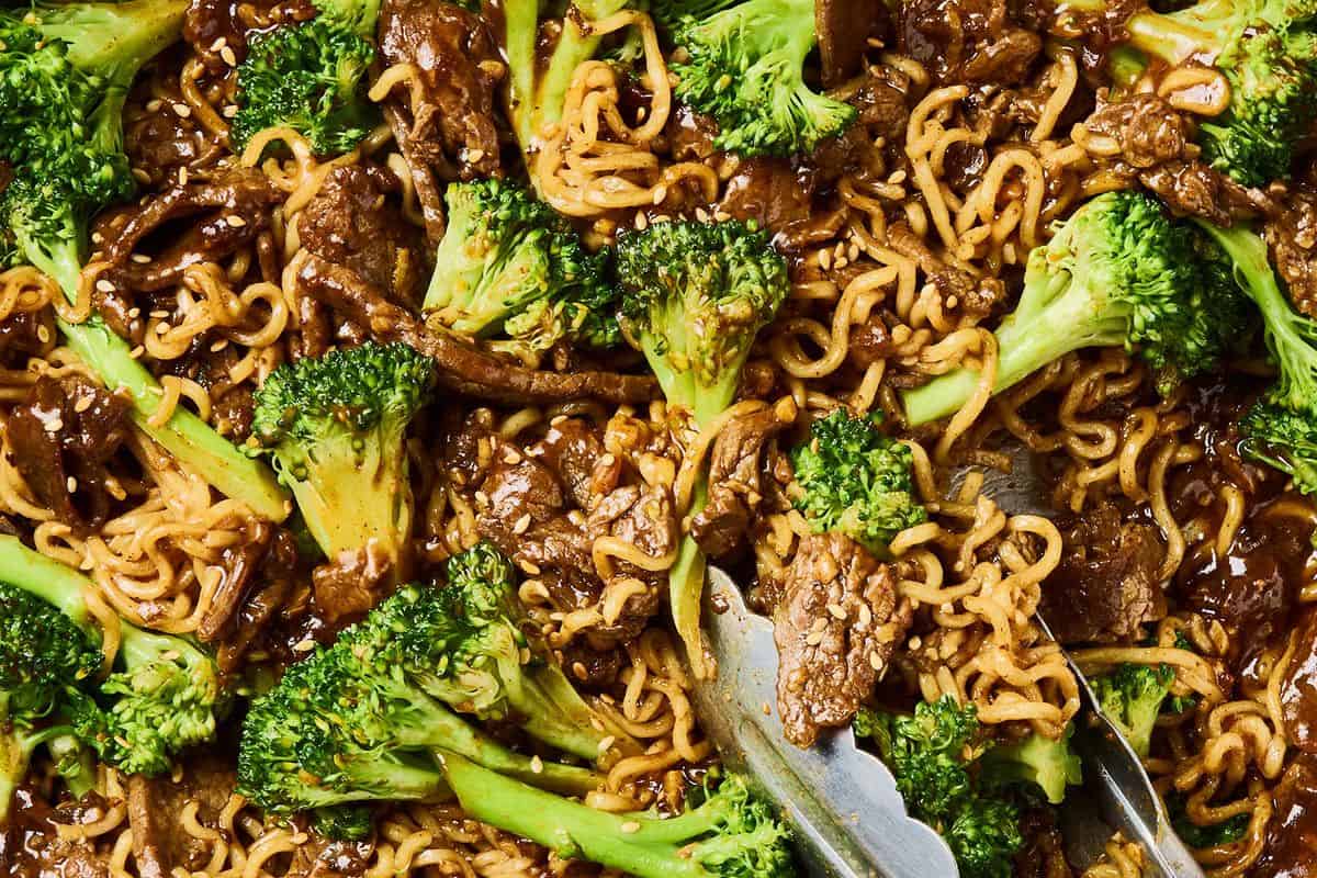 Close-up of flavor packed ramen with tender beef, fresh broccoli florets, and ramen noodles in a rich brown sauce, with a pair of metal tongs visible on the right side.