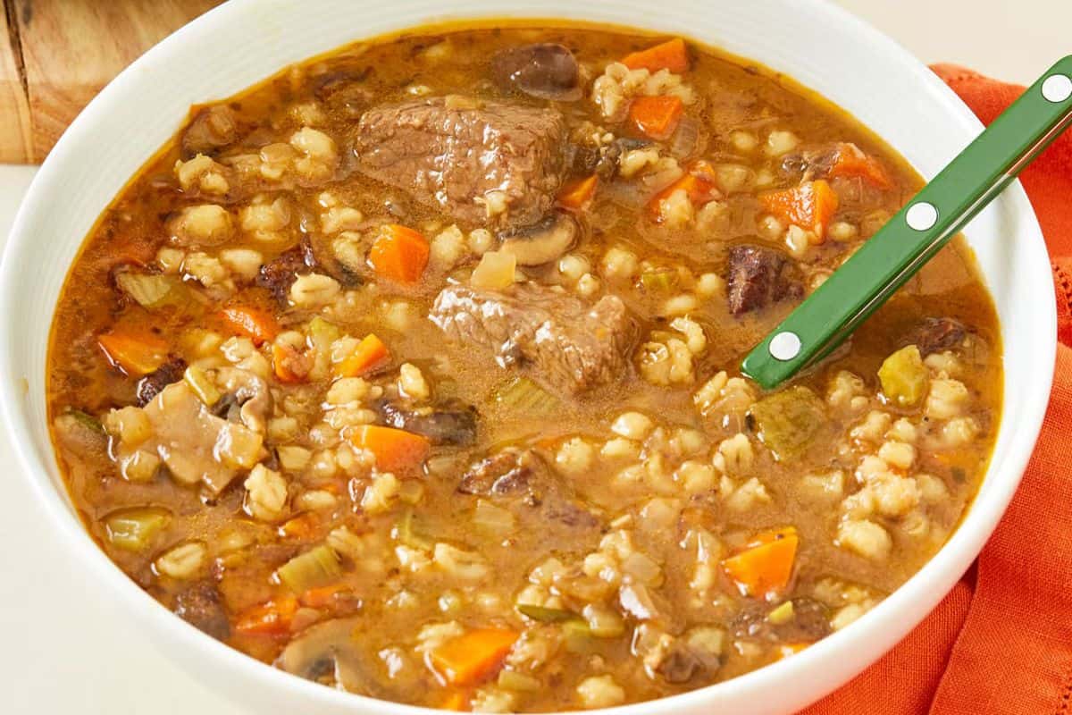 A bowl of comforting beef and barley soup with chunks of beef, carrots, celery, onions, mushrooms, and barley in a brown broth, served with a green-handled spoon—perfect for dinner.