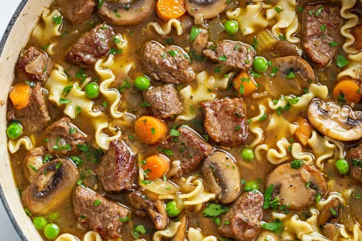 A close-up of beef noodle soup featuring chunks of beef, mushrooms, carrots, green peas, and curly pasta in a savory broth, garnished with chopped herbs—a comforting dinner perfect for your dinner rotation.