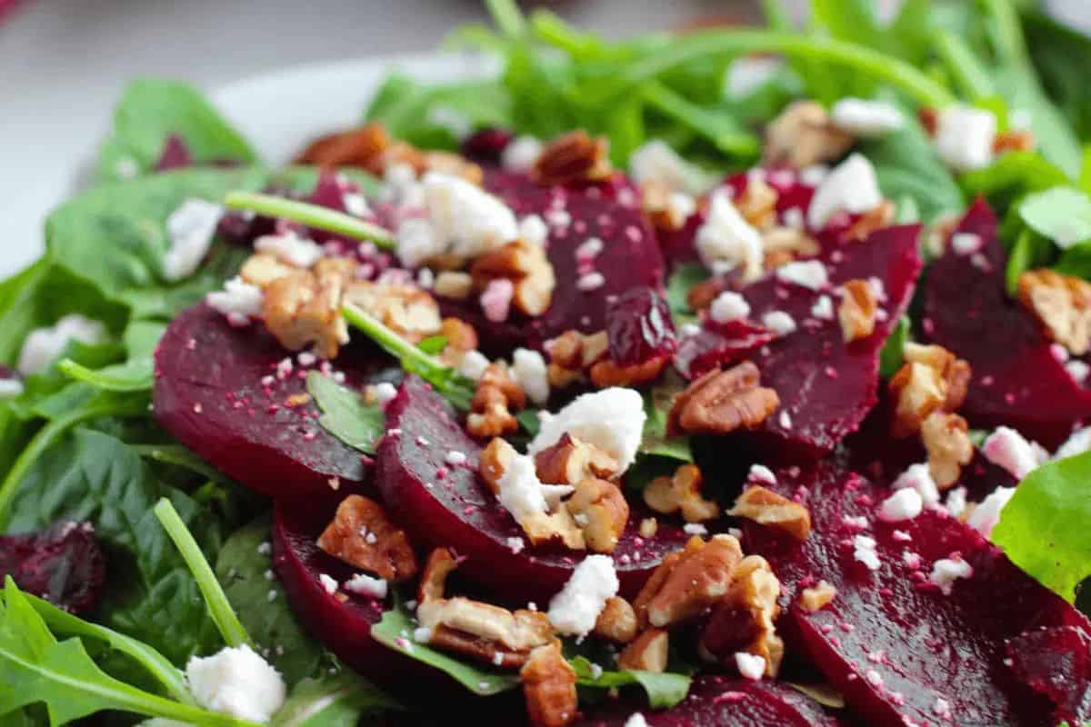 A close-up of a vibrant salad with sliced red beets, crumbles of white feta cheese, chopped pecans, and what appears to be arugula or mixed greens.