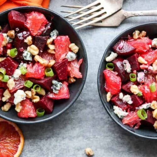 Two black bowls filled with a salad of beets, blood orange segments, walnuts, blue cheese crumbles, and sliced green onions on a gray surface—perfect inspiration for grapefruit recipes—served with forks and sliced oranges nearby.