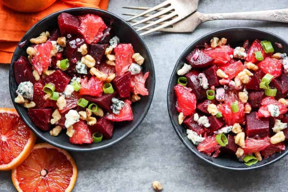Two black bowls filled with a salad of beets, blood orange segments, walnuts, blue cheese crumbles, and sliced green onions on a gray surface—perfect inspiration for grapefruit recipes—served with forks and sliced oranges nearby.