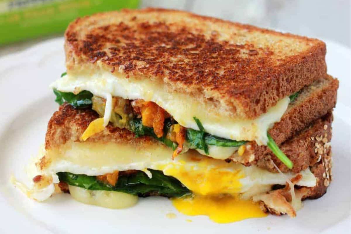 A grilled cheese sandwich with spinach, melted cheese, and a runny fried egg, cut in half and stacked on a white plate—one of our most delicious sandwiches.
