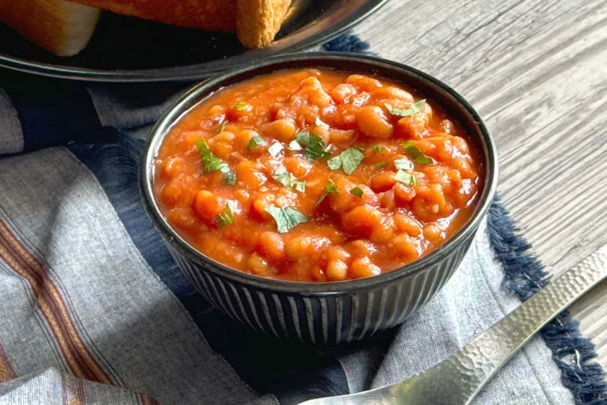 A bowl of canned beans in tomato sauce, garnished with chopped herbs, sits on a striped cloth with a spoon and toast in the background—perfect inspiration for delicious dishes and easy recipes.