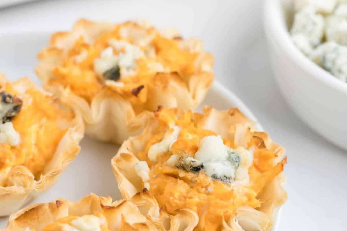 Mini phyllo pastry cups filled with a cheesy orange mixture and topped with crumbled blue cheese, served on a white plate—perfect for anyone looking for unique cold appetizer ideas.