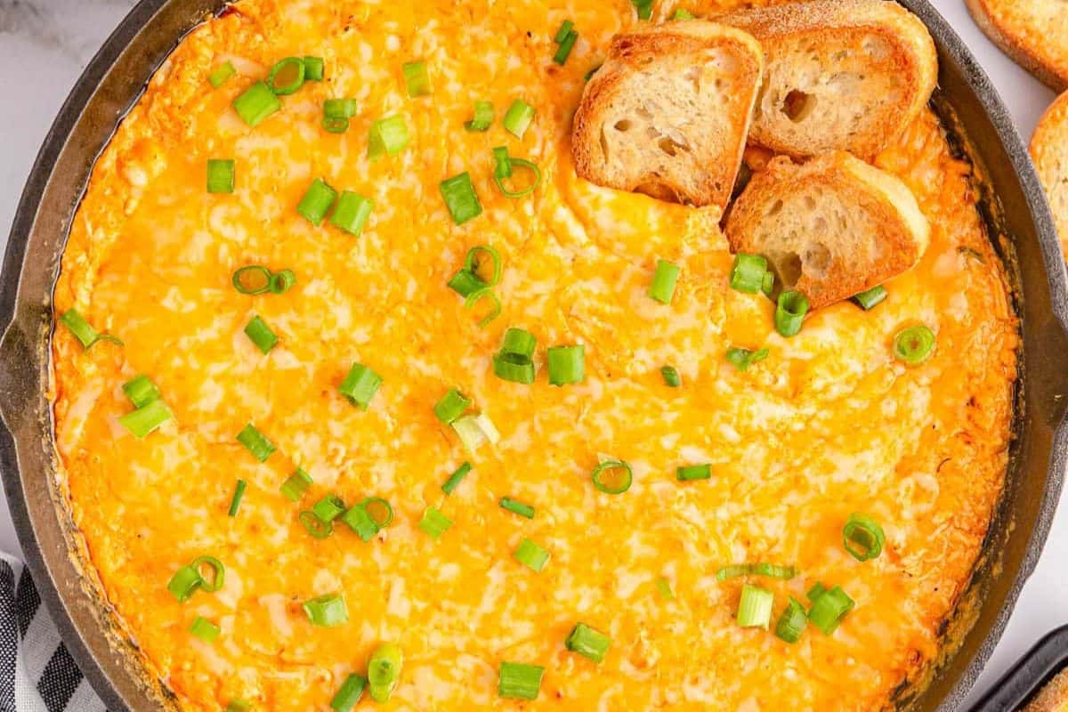 Baked cheesy dip topped with chopped green onions in a skillet, served with slices of toasted bread on the side—a crowd-pleaser perfect for party food recipes.