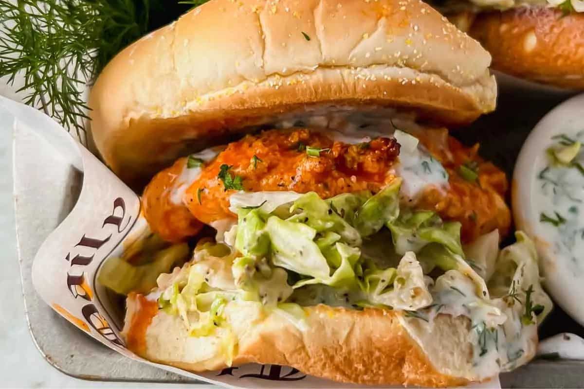A crispy chicken sandwich with lettuce and creamy sauce served in a bun, placed on parchment paper with a side of sauce nearby—a perfect choice for fans of gourmet sandwiches.
