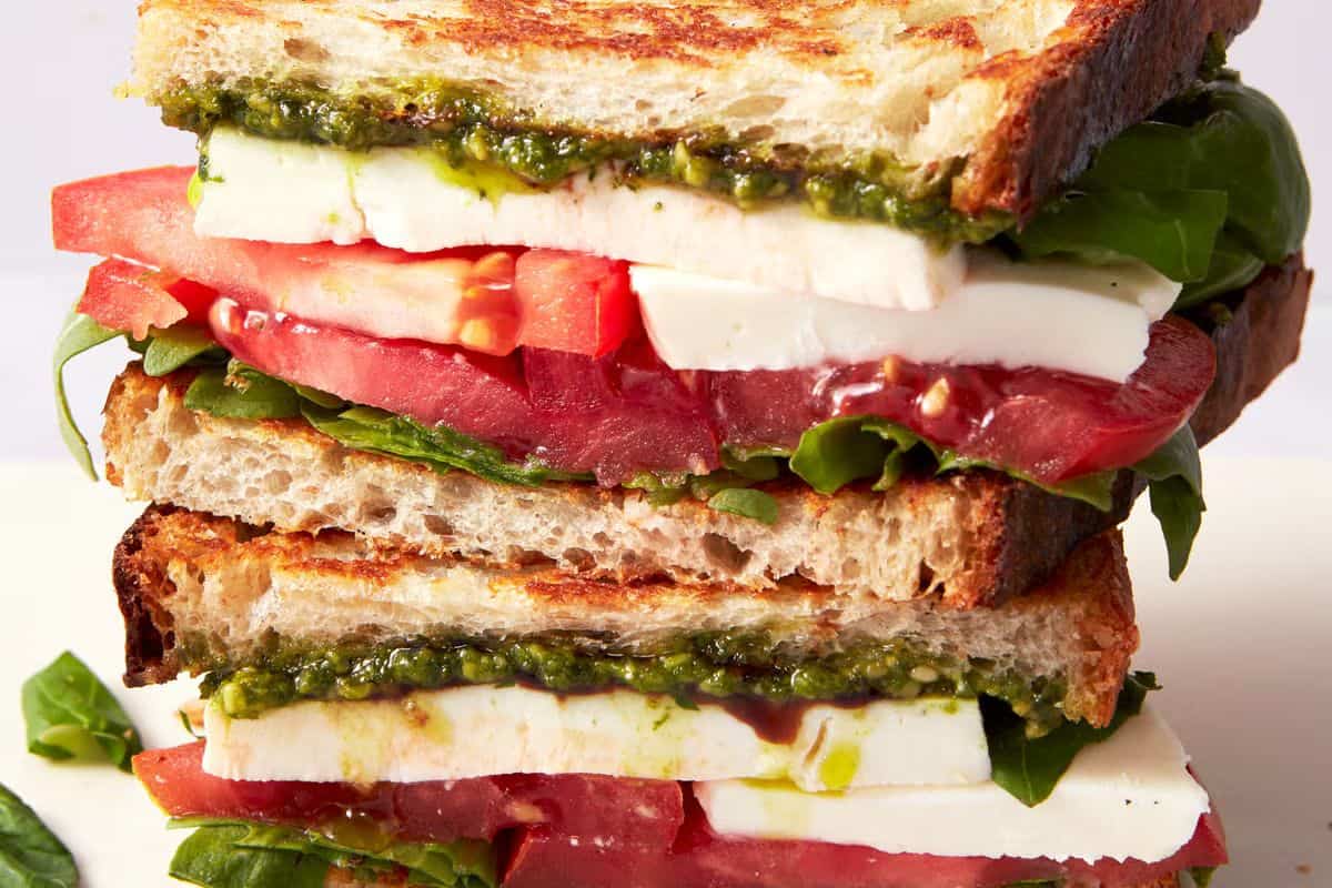 A close-up of two stacked gourmet sandwiches made with toasted bread, mozzarella cheese, tomato slices, fresh spinach, and a layer of pesto.