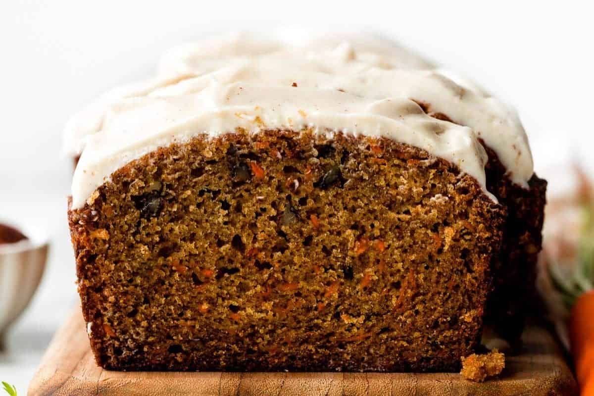 A close-up of a sliced loaf of carrot cake with white frosting on top, placed on a wooden board—a must try for fans of quick breads.